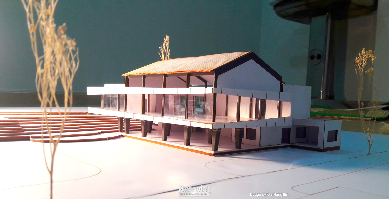 Mô hình kiến trúc cận cảnh hệ cột thép và vách kính khung đen của khối nhà, mái dốc phủ màu gỗ nhạt phía trên. Chi tiết kế…