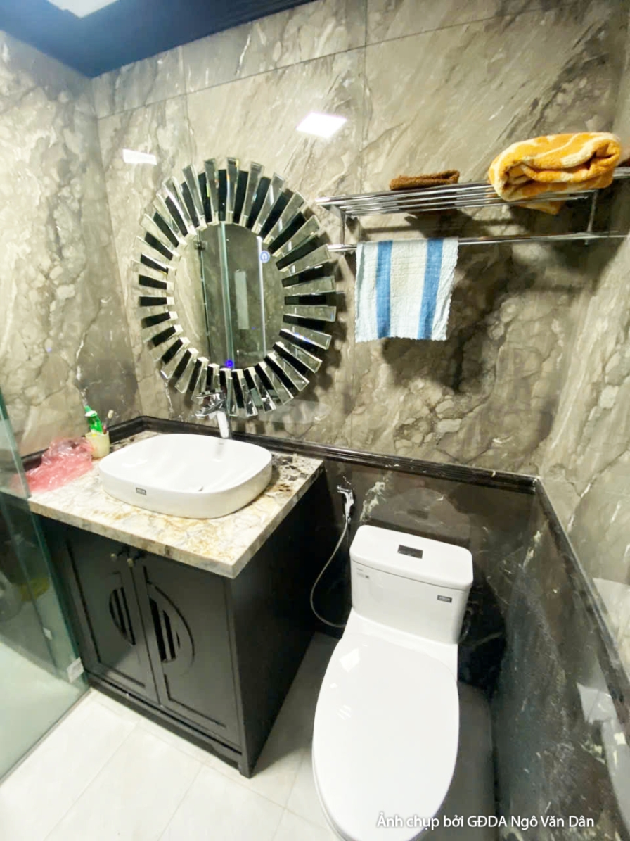 Phòng vệ sinh hiện đại với lavabo đặt trên tủ gỗ tối màu, gương tròn lớn và vách kính tắm đứng. Tường ốp đá vân tối màu, t…