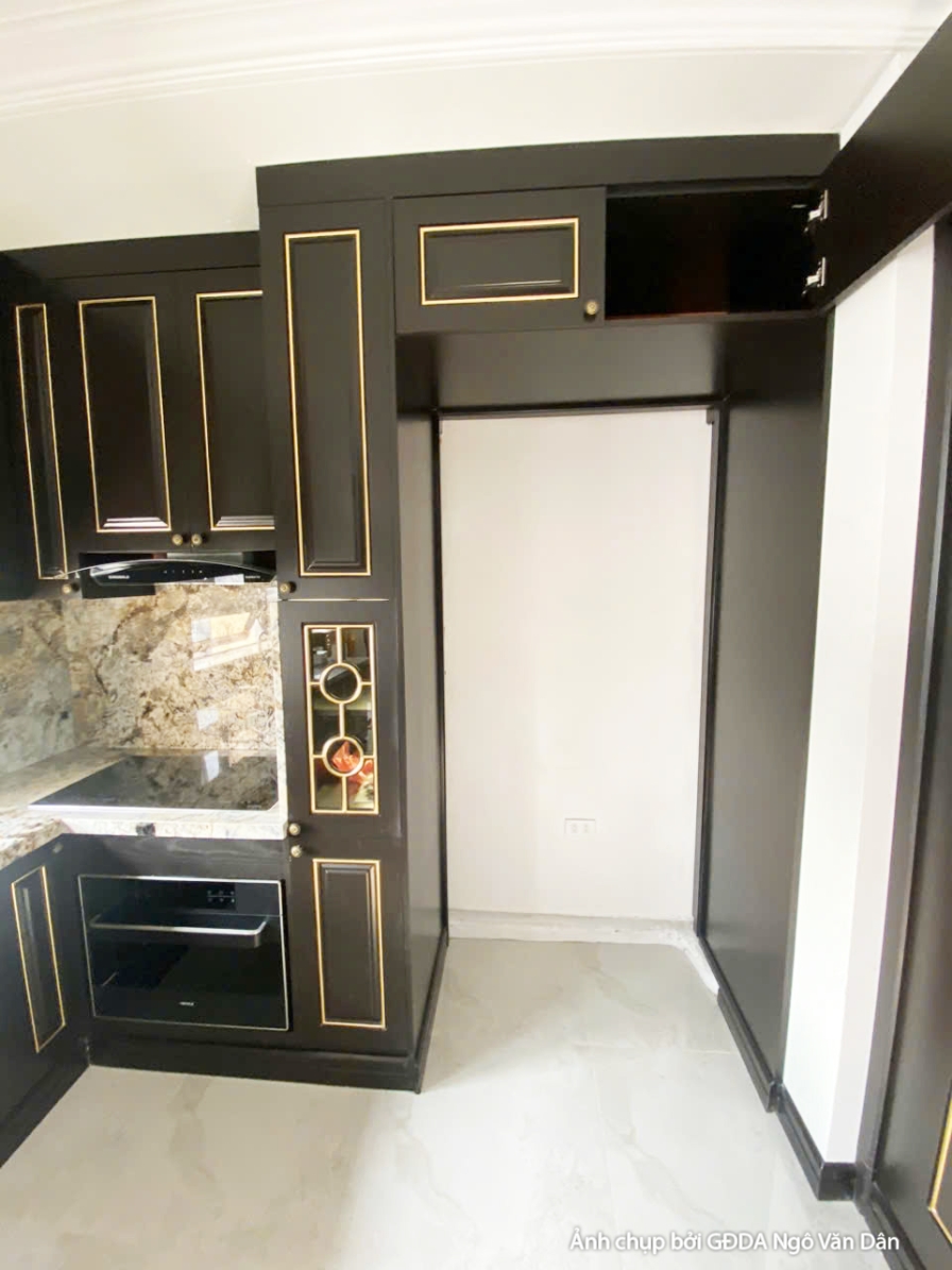 Khu vực bếp hiện đại với hệ tủ bếp trên dưới màu đen, mặt bàn đá sáng màu và bồn rửa đôi. Bố cục gọn gàng, tối ưu công năn…