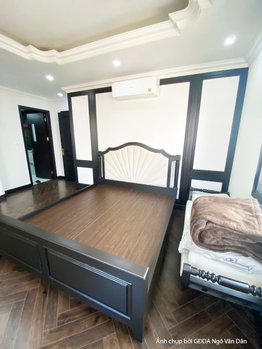 Phòng ngủ với giường gỗ tối màu, tủ đầu giường và cửa sổ lớn. Rèm cửa màu trung tính, không gian đang được kiểm tra kỹ lưỡ…