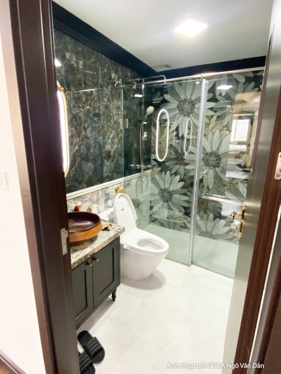Phòng vệ sinh hiện đại với lavabo đặt trên tủ gỗ tối màu, gương tròn lớn và vách kính tắm đứng. Tường ốp đá vân tối màu, t…