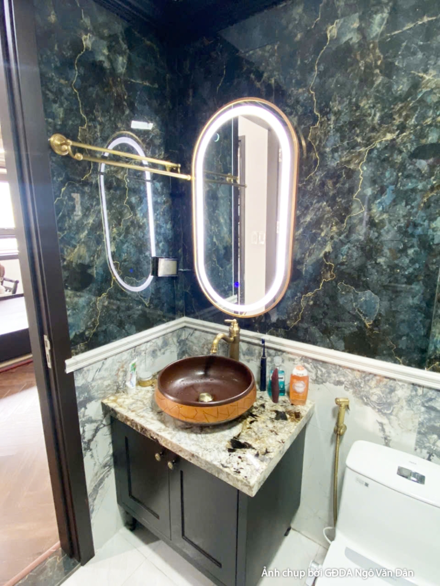 Phòng vệ sinh hiện đại với lavabo đặt trên tủ gỗ tối màu, gương tròn lớn và vách kính tắm đứng. Ánh sáng từ đèn trần và gư…