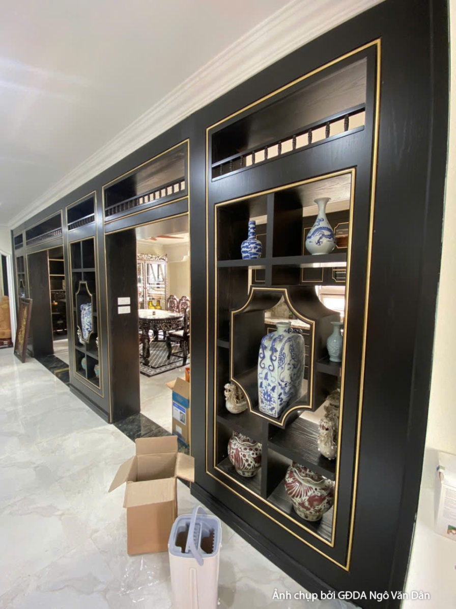 Phòng khách với bộ bàn ghế gỗ chạm khắc tinh xảo, kết hợp tủ kệ trưng bày đồ trang trí. Sàn gạch hoa văn và trần giật cấp…