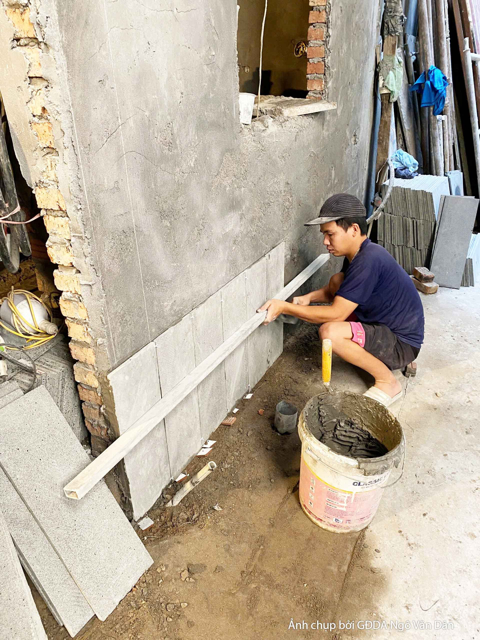 Công nhân đang tỉ mỉ ốp từng viên gạch men lên bề mặt tường trong không gian nội thất. Bố cục cận cảnh, tập trung vào kỹ t…