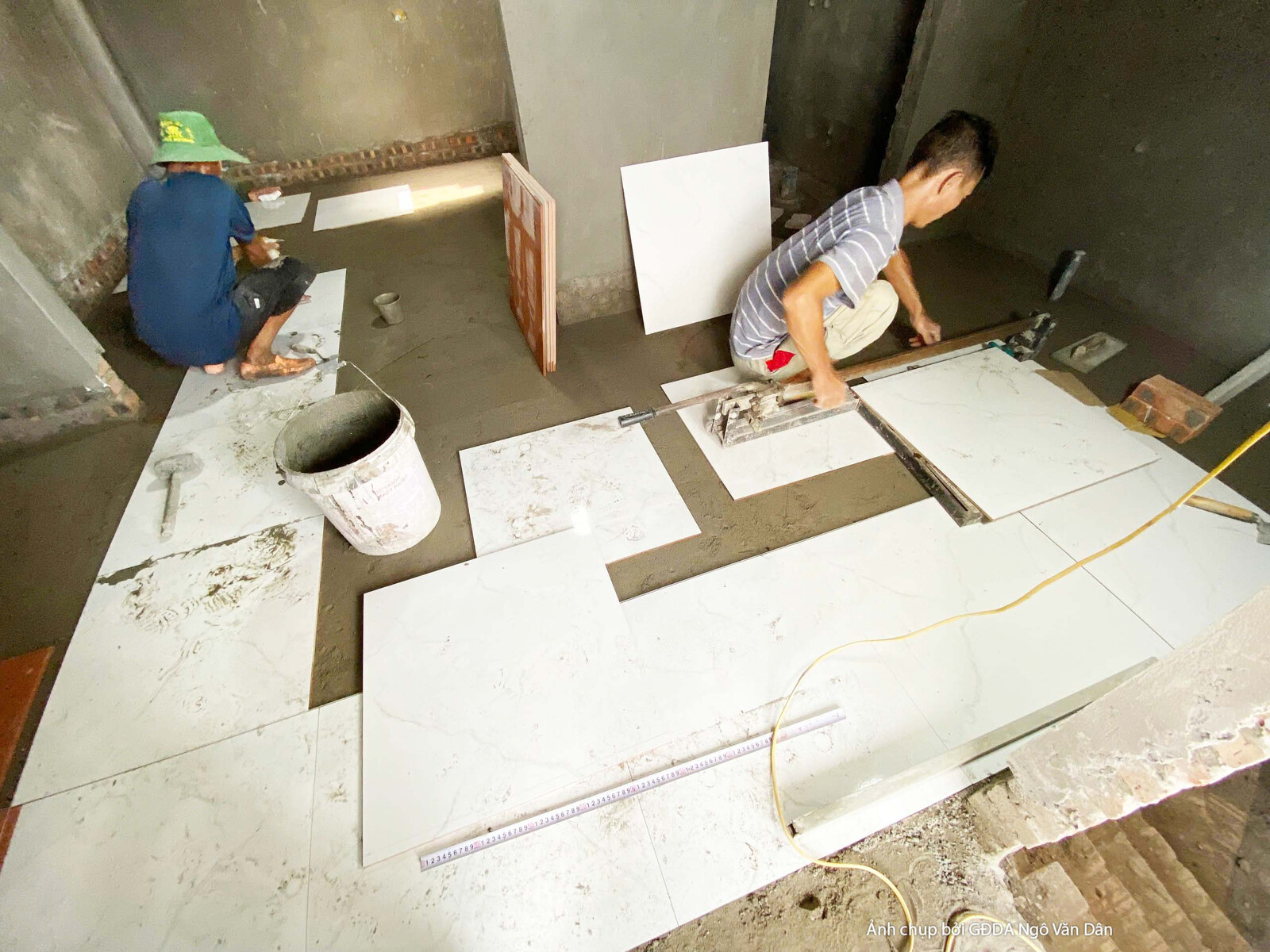 Công nhân đang tỉ mỉ lát những viên gạch men khổ lớn trên sàn nhà, sử dụng keo dán gạch và dụng cụ chuyên dụng. Bố cục cận…