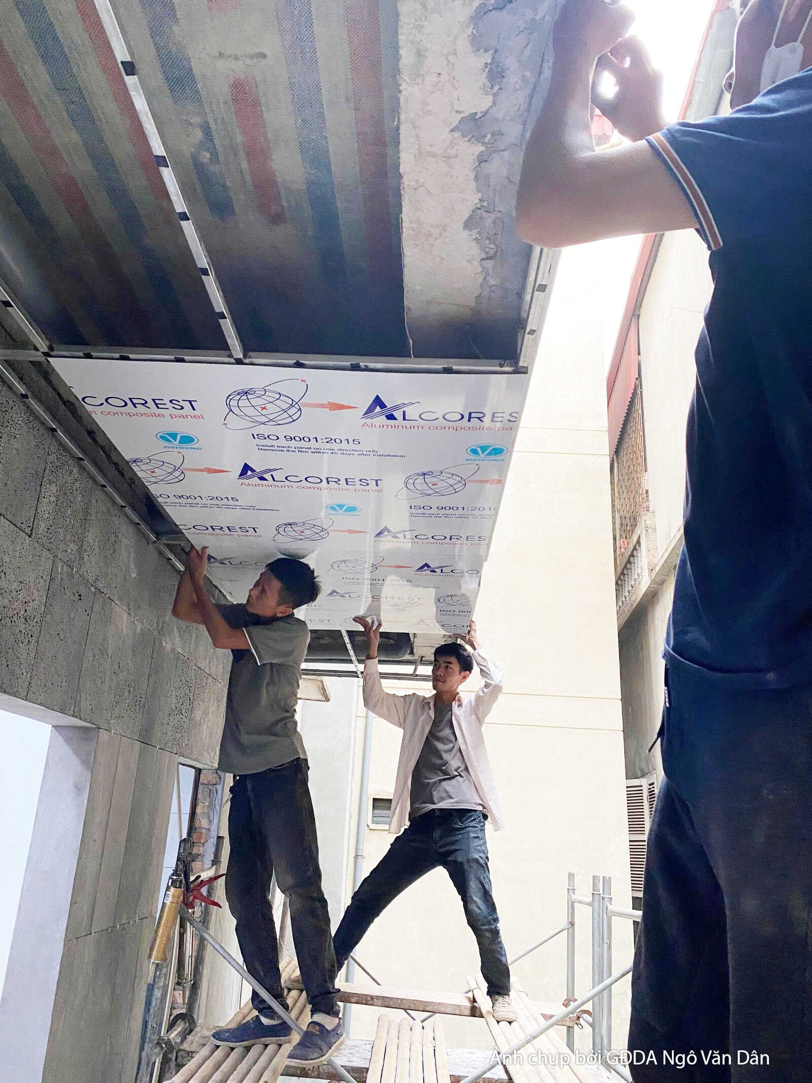 Hai công nhân đang lắp đặt hệ thống trần hoặc mái che, sử dụng các tấm vật liệu nhẹ và khung kim loại. Bố cục từ dưới lên…