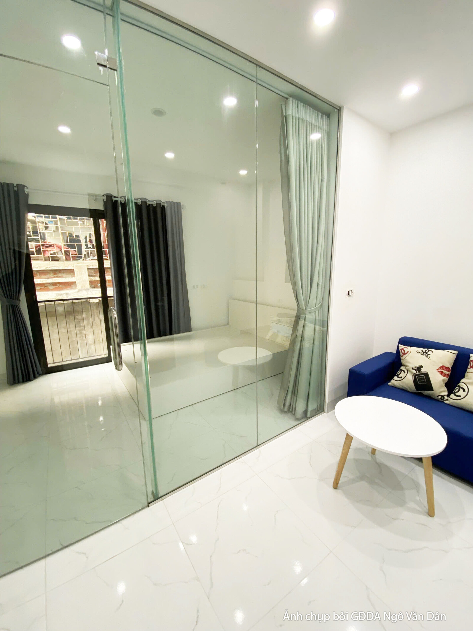 Không gian phòng khách nhỏ gọn, hiện đại với bộ sofa màu xanh dương nổi bật, bàn trà tròn trắng và vách kính cường lực tro…