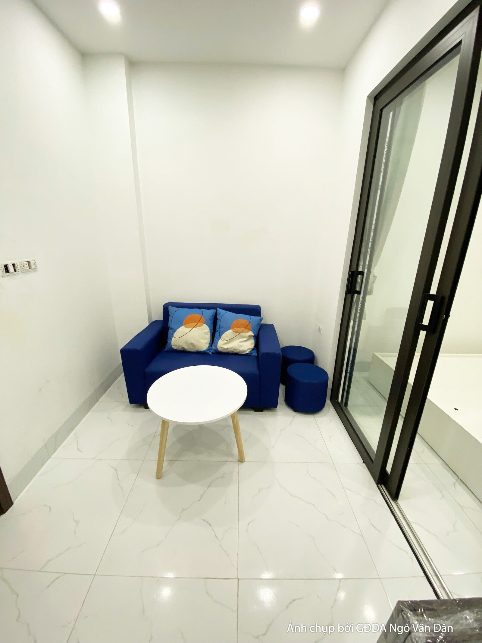 Góc nhìn từ cửa vào phòng khách, tập trung vào bộ sofa màu xanh và bàn trà tròn. Cửa kính lớn với rèm vải xám giúp điều ch…