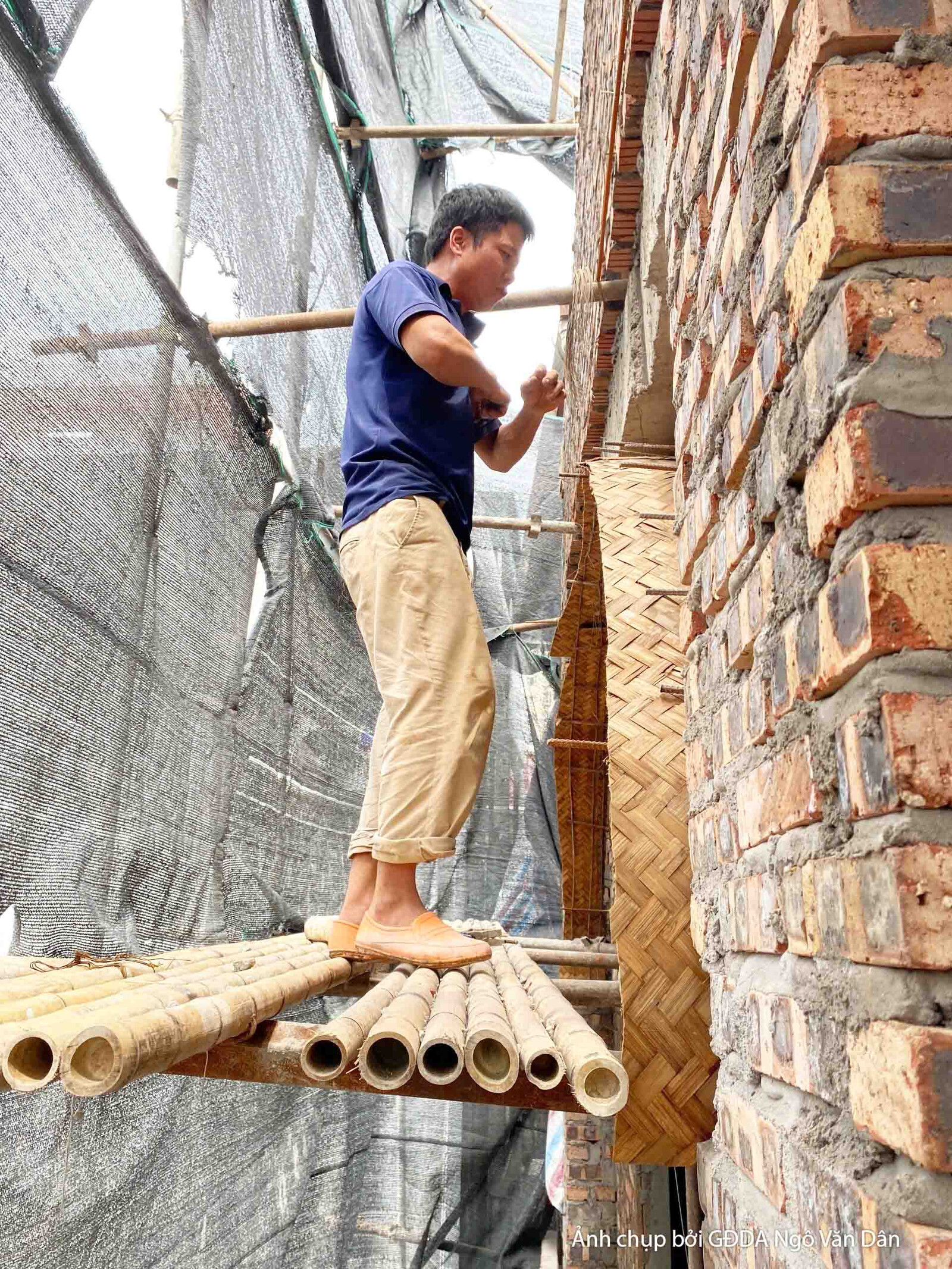 Công nhân đang ốp gạch hoặc vật liệu trang trí lên bề mặt tường ngoại thất, đứng trên giàn giáo tre truyền thống. Bố cục c…