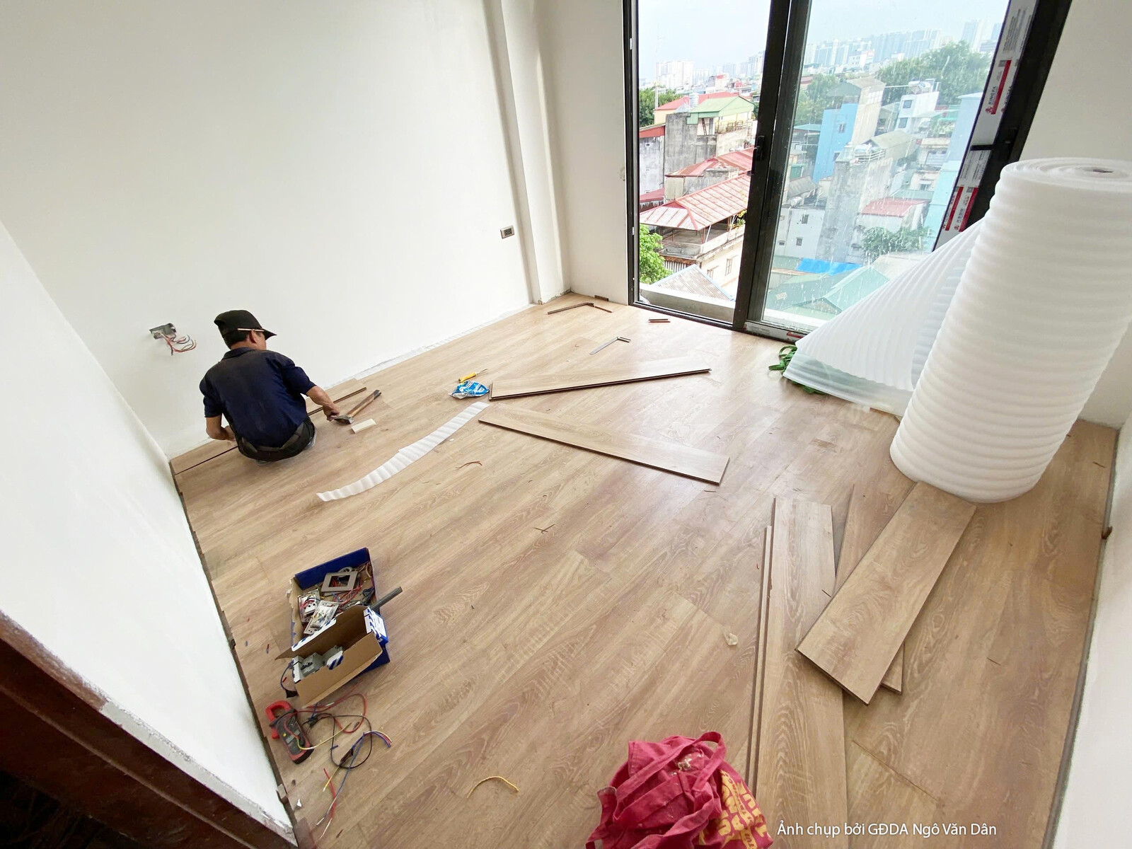 Không gian phòng ngủ đang trong giai đoạn lắp đặt sàn gỗ công nghiệp. Bố cục từ góc nhìn ngang cho thấy các tấm sàn gỗ màu…