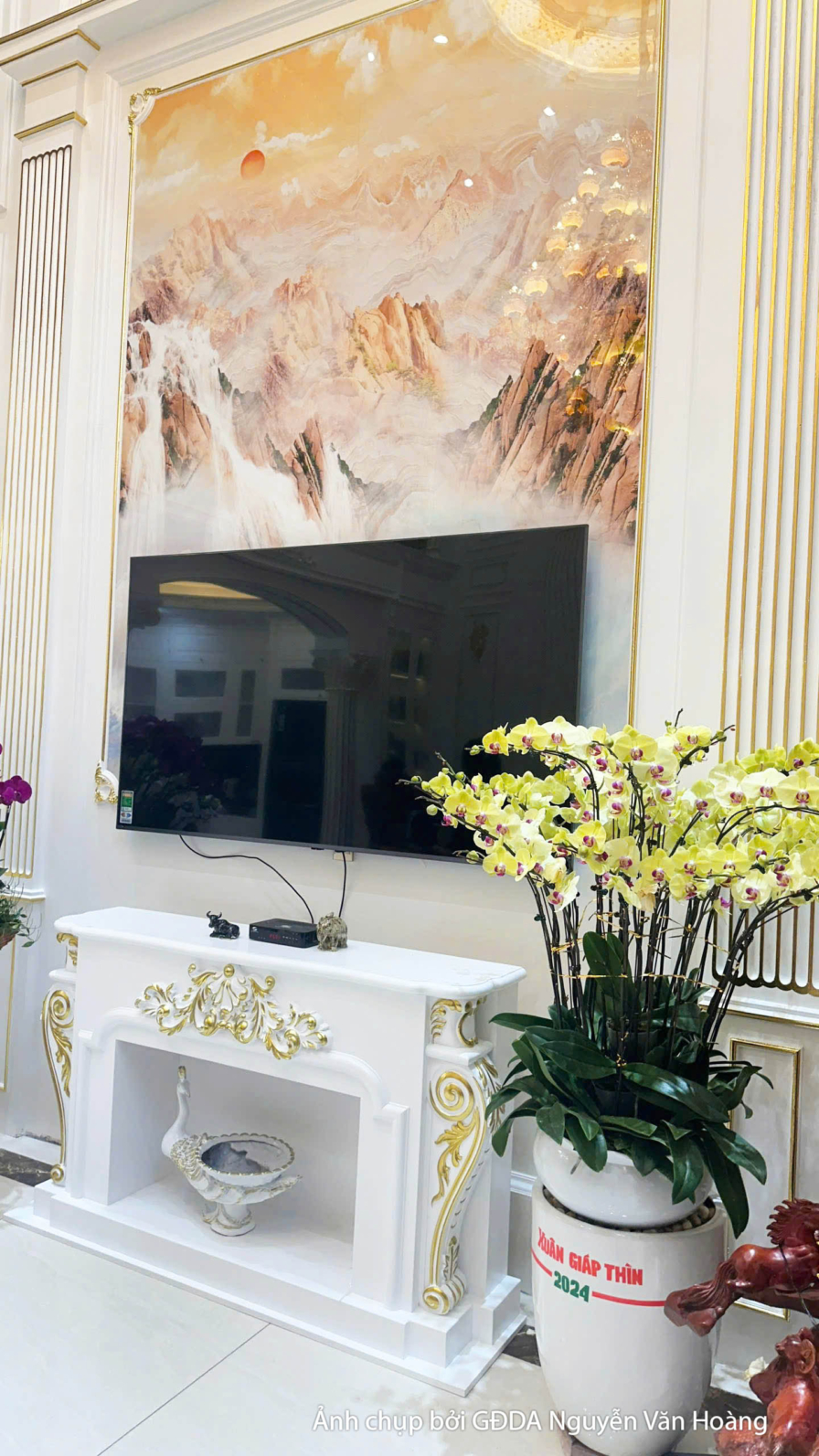 Cận cảnh khu vực kệ tivi và vách trang trí phòng khách. Bức tranh nghệ thuật lớn với tông màu ấm áp, kết hợp bình hoa tran…