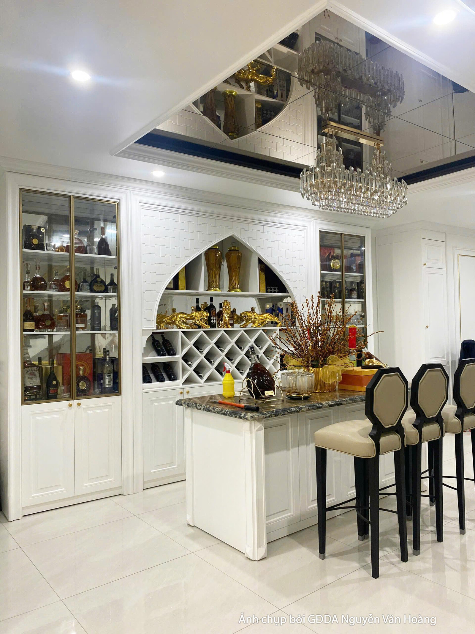 Toàn cảnh khu vực bếp và quầy bar đã hoàn thiện. Tủ bếp màu trắng tinh tế, đảo bếp lớn với mặt đá đen, kết hợp đèn chùm ph…