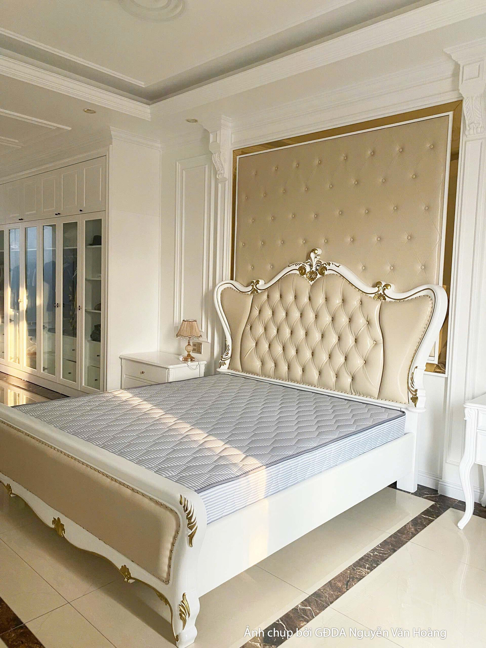 Cận cảnh giường ngủ bọc nệm màu kem với đầu giường cao, thiết kế tân cổ điển. Đèn ngủ treo tường hai bên và các chi tiết p…
