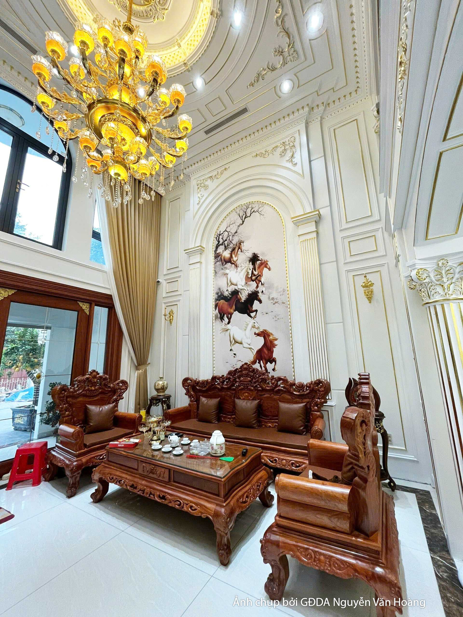 Phòng khách đã hoàn thiện, góc nhìn từ dưới lên, làm nổi bật chiều cao thông tầng và đèn chùm pha lê. Cầu thang với lan ca…