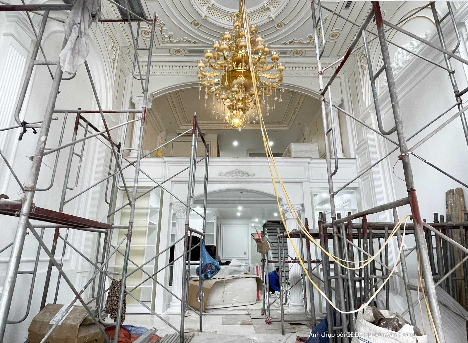 Sảnh thông tầng đang trong giai đoạn lắp đặt hoàn thiện, với giàn giáo cao phục vụ việc thi công trần và tường. Đèn chùm p…