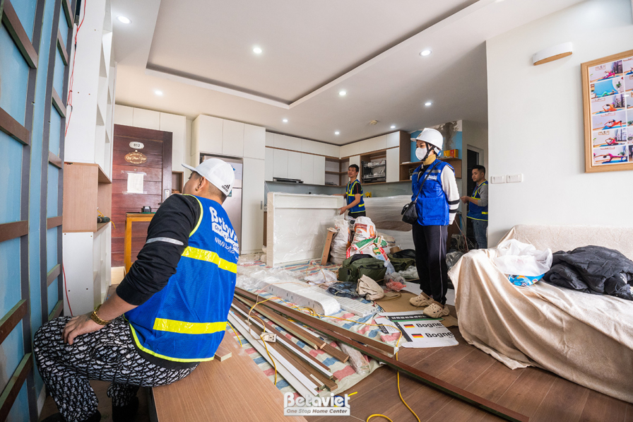Cận cảnh phòng khách đang lắp đặt nội thất. Thành viên Betaviet kiểm tra chi tiết tấm ốp tường và kệ gỗ. Không gian tông m…