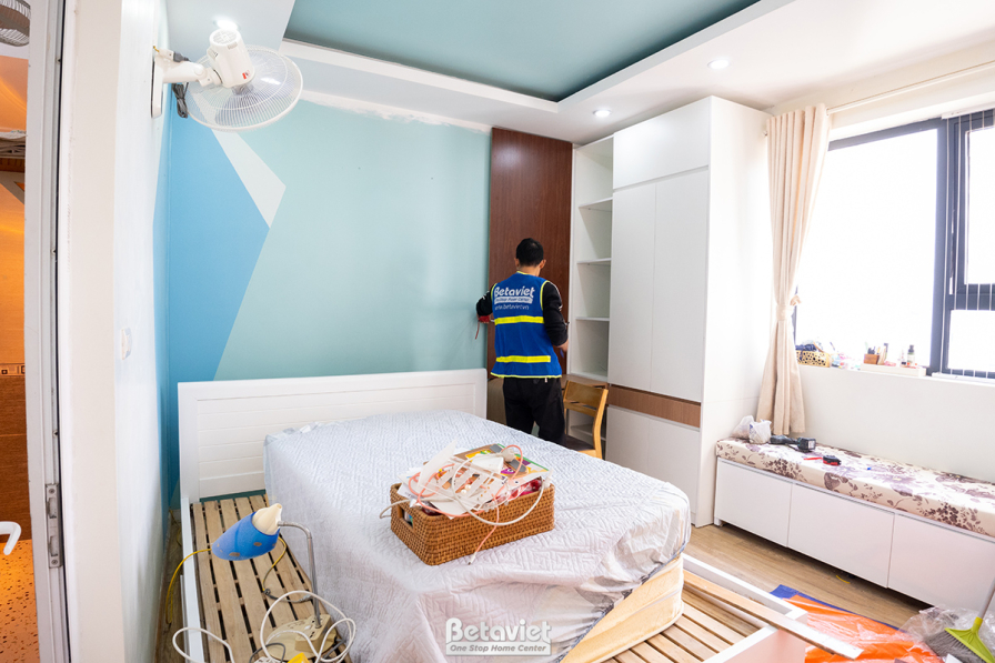 Phòng ngủ đang trong giai đoạn lắp đặt hoàn thiện, với tủ quần áo lớn màu trắng và vách đầu giường ốp màu xanh pastel. Ánh…