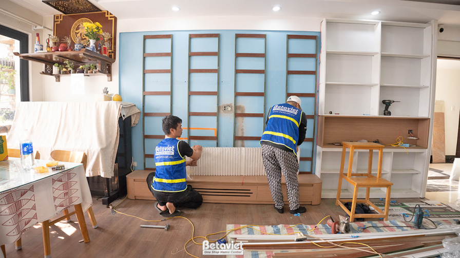 Góc nhìn tổng thể phòng khách và bếp đang hoàn thiện. Tủ kệ màu trắng và xanh pastel được lắp đặt, tạo điểm nhấn tươi sáng…