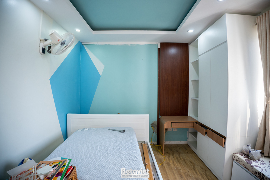 Góc nhìn tổng thể một phòng ngủ với giường ngủ đơn giản, tủ quần áo âm tường màu nâu gỗ và vách đầu giường ốp màu xanh pas…