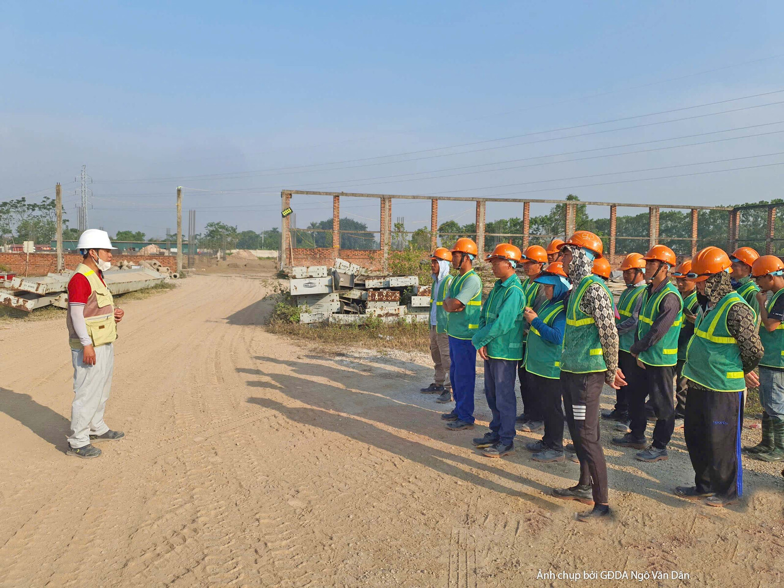 Nhóm kỹ sư và công nhân đang họp giao ban tại công trường, đứng trên nền đất trống. Bố cục cho thấy sự trao đổi công việc…