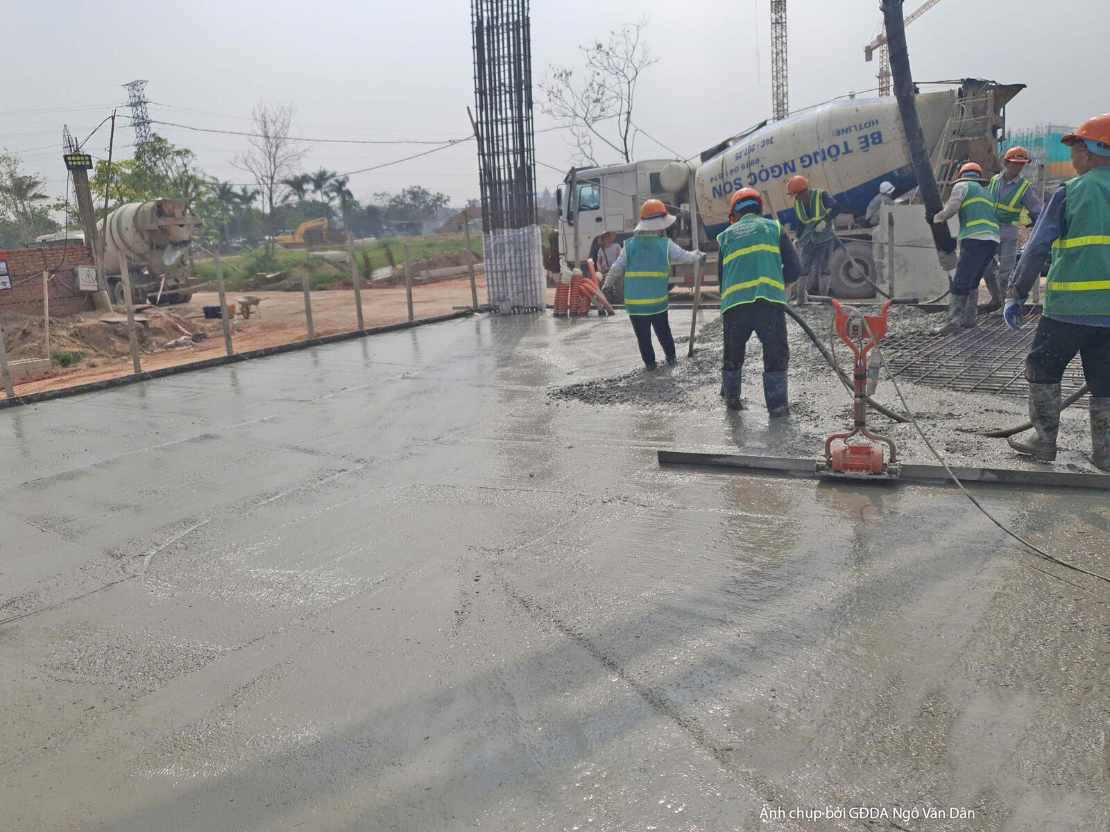 Công nhân đang thực hiện đổ bê tông cho sàn, sử dụng xe bồn và các dụng cụ san gạt. Bố cục rộng, cho thấy quá trình đổ bê…