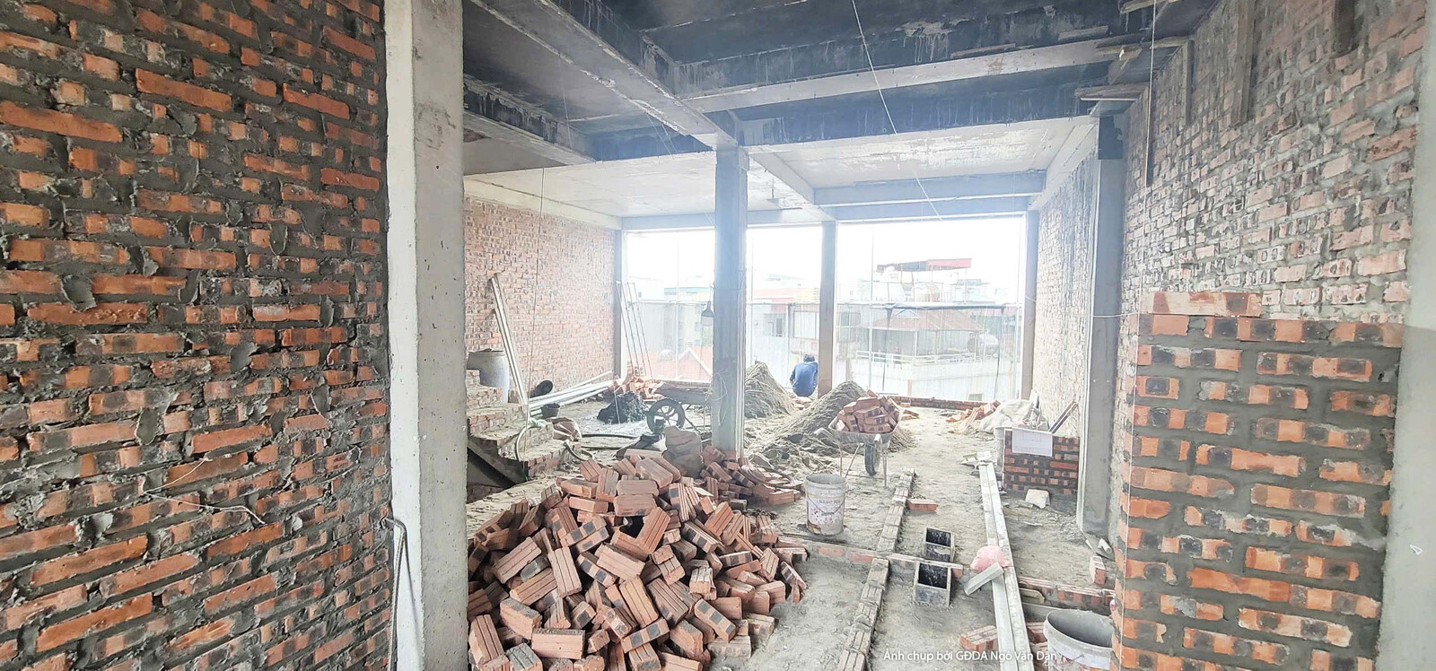 Một không gian nội thất khác đang trong giai đoạn thi công phần thô, với các bức tường gạch đỏ cam chưa trát. Cửa sổ lớn c…