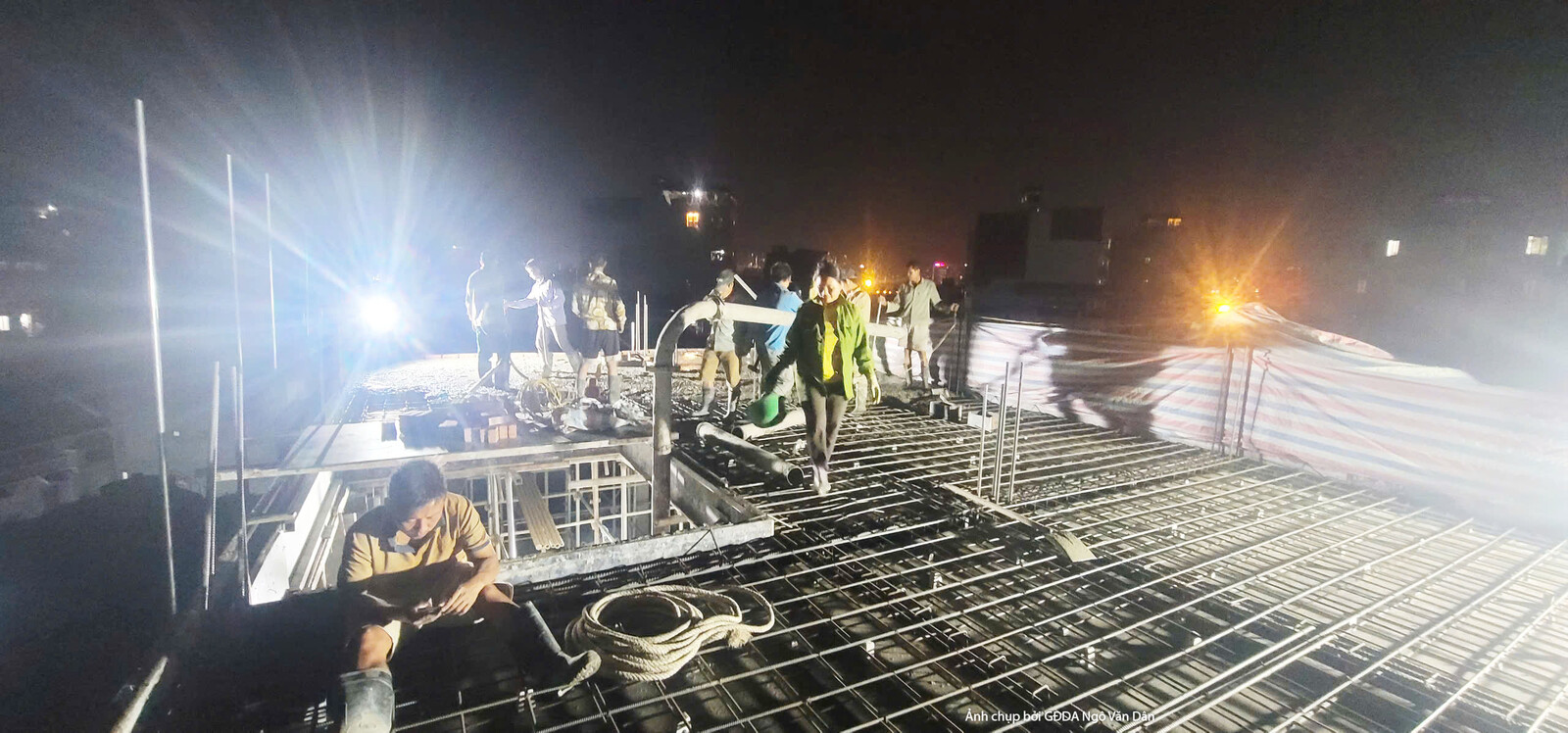Cảnh công nhân đang làm việc khẩn trương trên sàn bê tông cốt thép vào ban đêm, dưới ánh đèn chiếu sáng mạnh mẽ. Điều này…