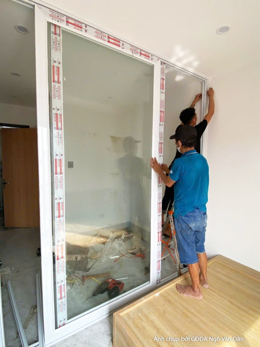 Không gian ban công đang lắp đặt cửa kính trượt lớn, nhìn từ trong phòng ra. Hai công nhân phối hợp dựng khung nhôm kính c…
