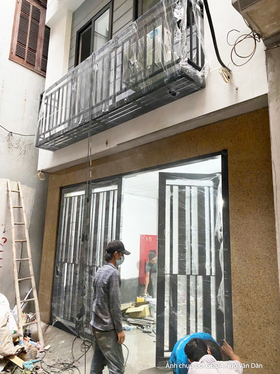 Công nhân đang lắp đặt khung cửa sổ lớn bằng nhôm kính tại mặt tiền công trình. Khung cửa màu đen tạo điểm nhấn trên nền t…