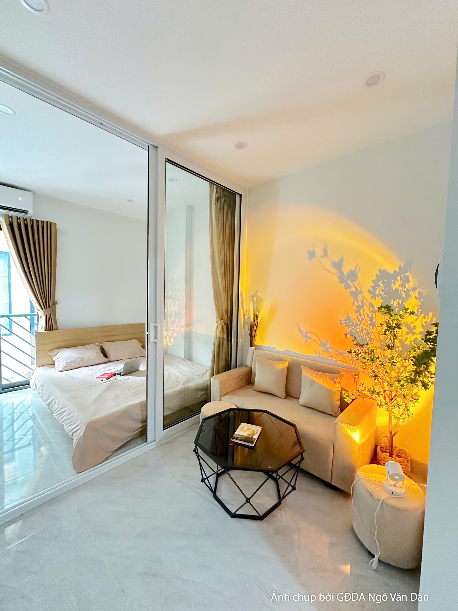 Không gian phòng ngủ nhìn qua vách kính trượt sang khu vực phòng khách của căn hộ hoàn thiện. Giường nệm màu be, tủ gỗ sán…