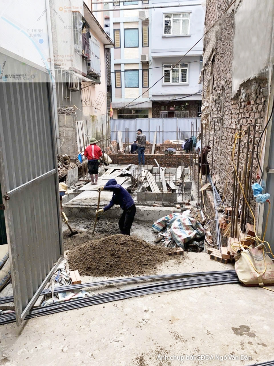 Công trường thi công phần thô, nhìn từ góc đường hẹp, có công nhân đang làm việc. Các vật liệu xây dựng như cát, đá, sắt t…
