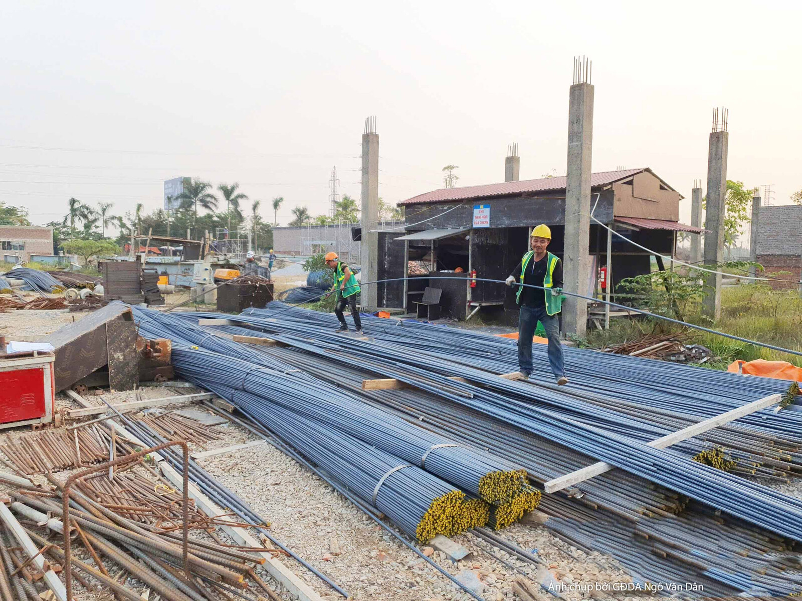 Khu vực tập kết vật liệu xây dựng tại công trường. Các bó thép lớn được sắp xếp gọn gàng trên nền đất, một công nhân đang…