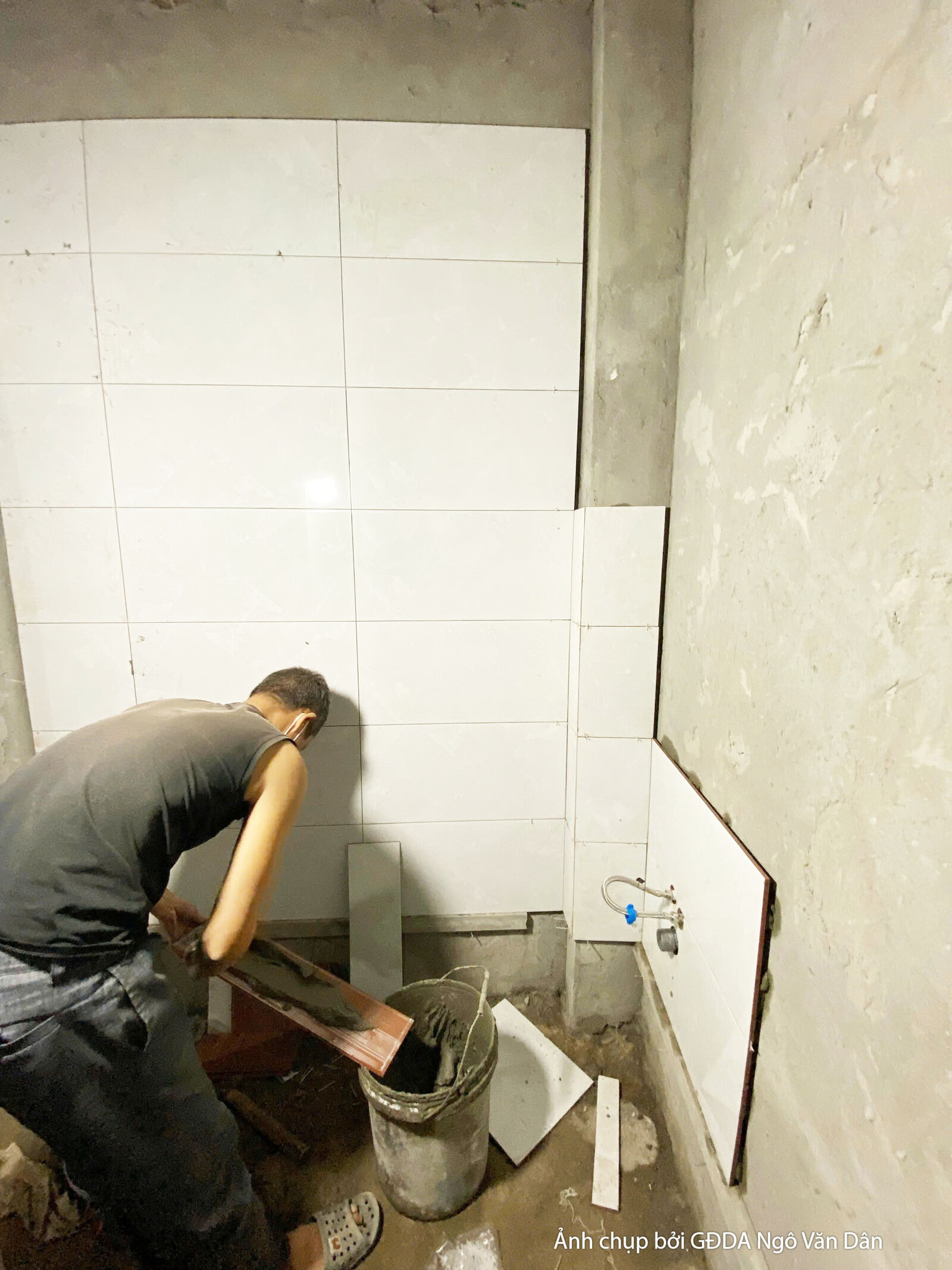 Công nhân đang ốp gạch men màu trắng lên tường trong không gian nội thất, có thể là khu vực bếp hoặc vệ sinh. Các đường mạ…