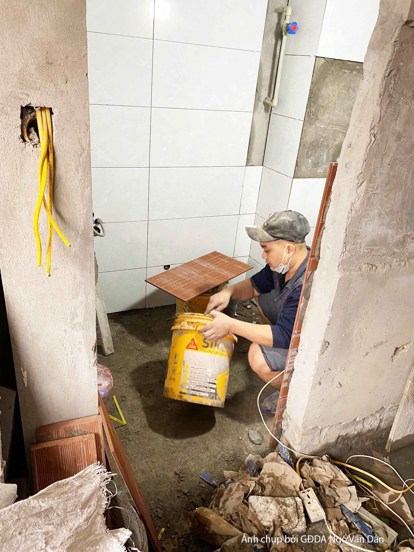 Cận cảnh công nhân đang ốp gạch tường, sử dụng keo dán gạch chuyên dụng. Bề mặt tường đã được ốp một phần, cho thấy sự tỉ…