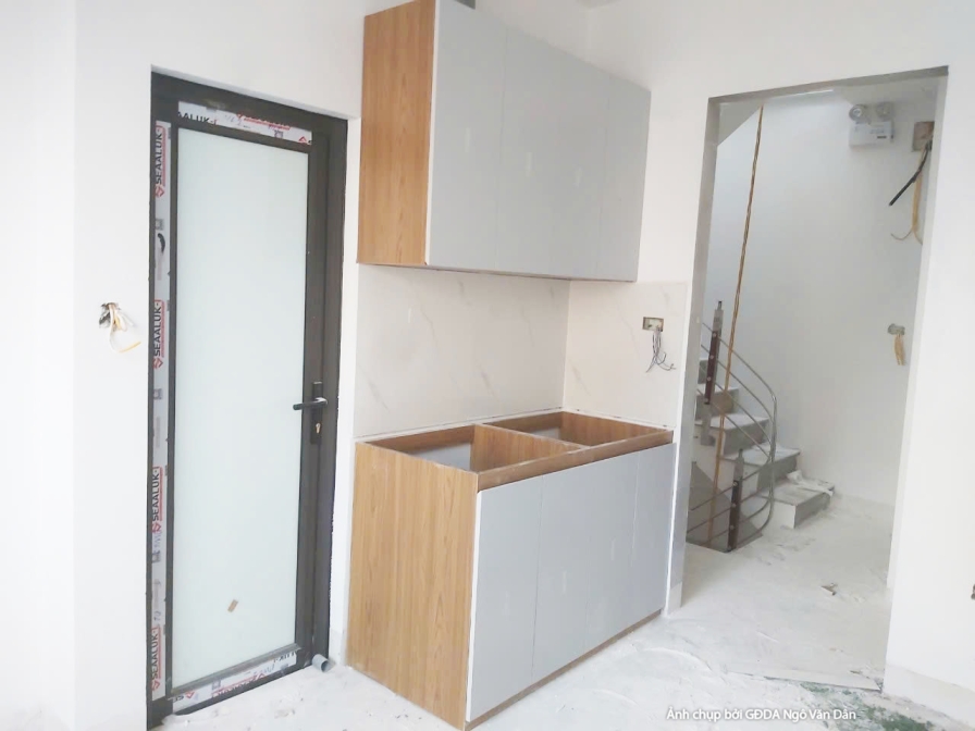 Khu vực bếp đang trong giai đoạn lắp đặt tủ bếp trên và dưới. Tủ bếp dưới màu vân gỗ sáng, tủ bếp trên màu trắng, thiết kế…
