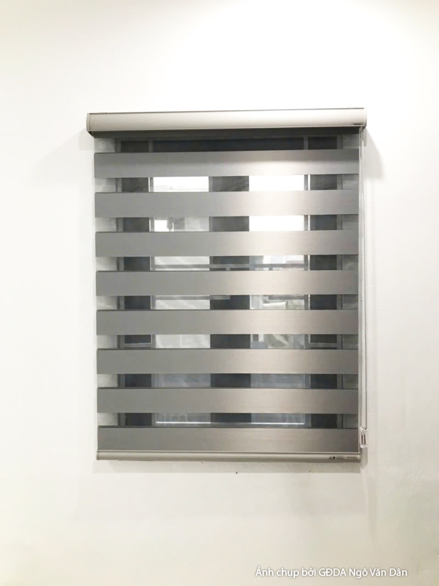 Cận cảnh rèm cuốn ngang màu xám bạc với các sọc ngang, được lắp đặt trên cửa sổ. Rèm có khả năng điều chỉnh ánh sáng linh…