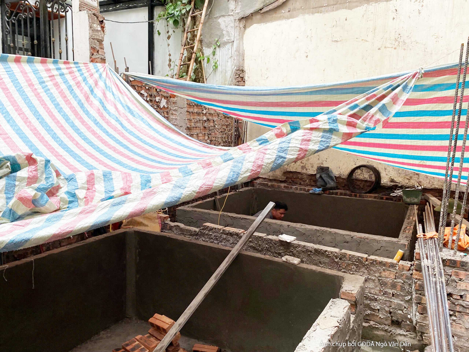 Khu vực hố móng hoặc bể ngầm đang được che chắn bằng bạt sọc xanh trắng. Bên trong có các thanh chống và vật liệu xây dựng…