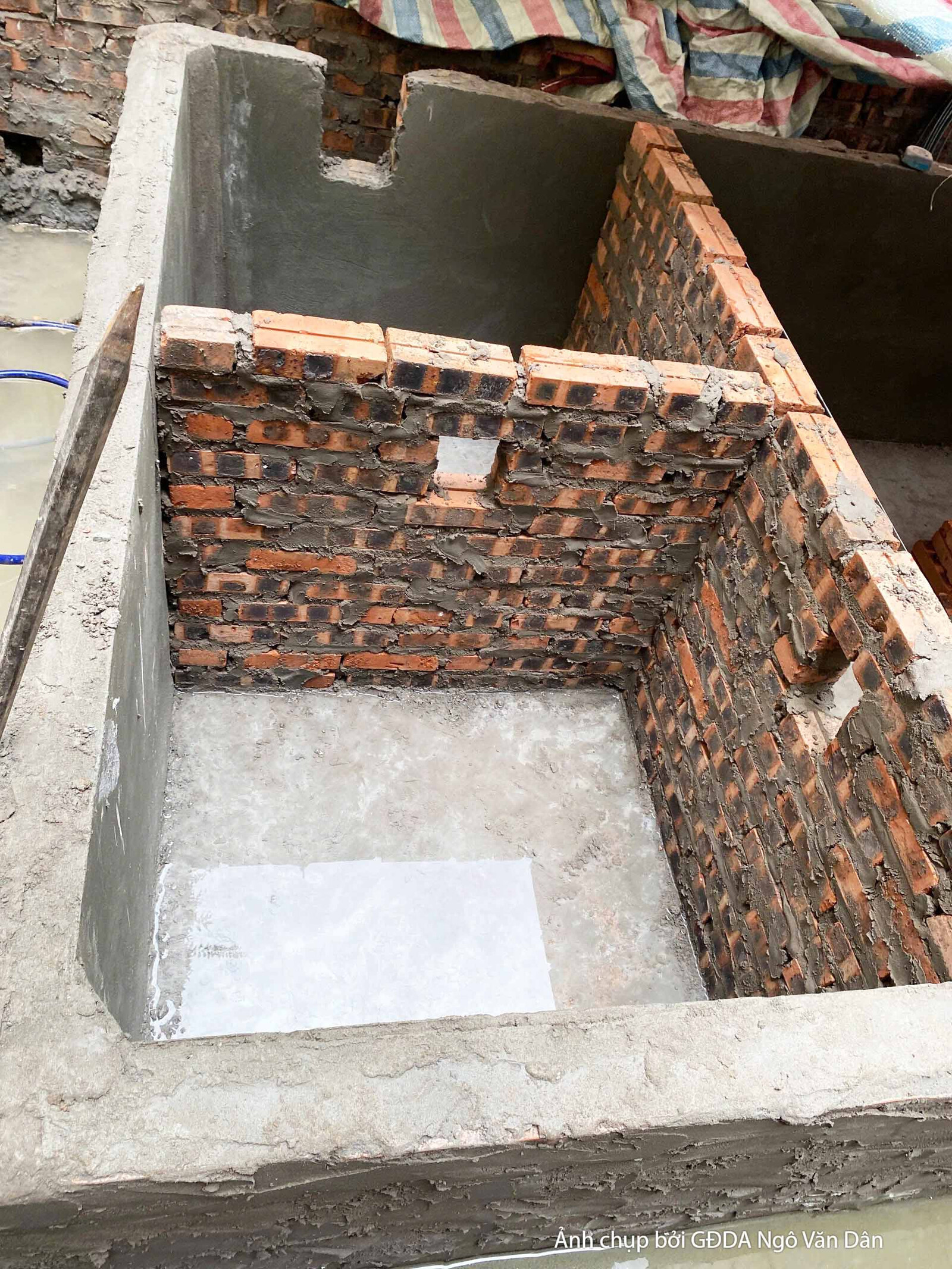 Cận cảnh một hố móng hoặc bể ngầm đã được xây tường gạch bao quanh, phần đáy được đổ bê tông phẳng. Đây là một phần của kế…