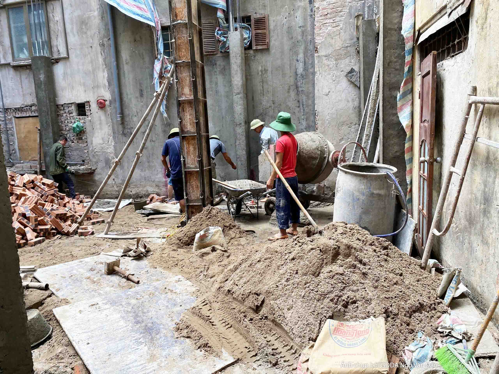 Công nhân đang trộn vữa hoặc bê tông thủ công bằng máy trộn và xẻng tại công trường. Xung quanh là đống cát, đá và xi măng…
