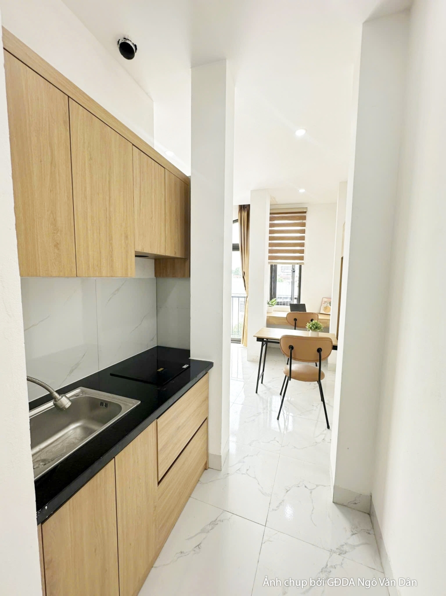 Khu vực bếp nhỏ gọn và hiện đại với tủ bếp trên và dưới bằng gỗ công nghiệp màu sáng, mặt bếp ốp đá đen. Sàn lát gạch vân…