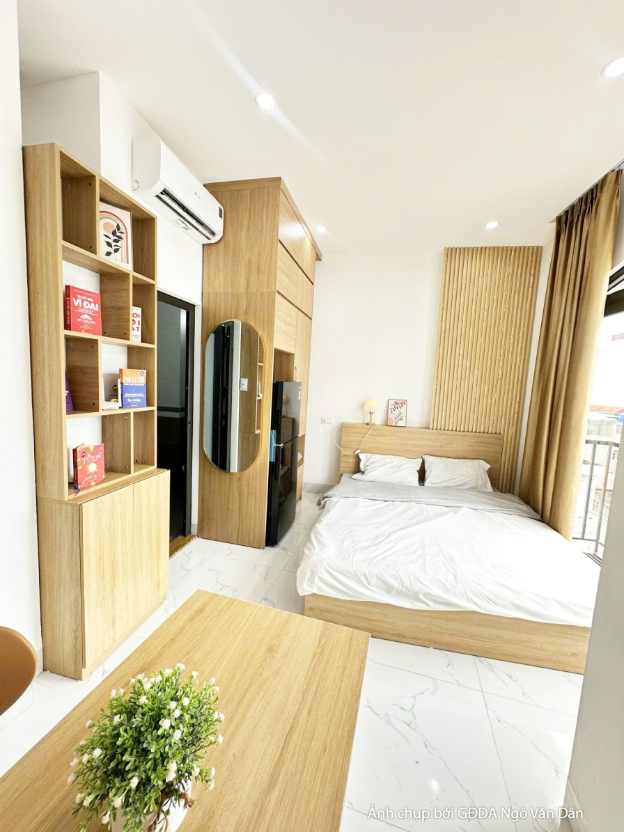 Góc phòng ngủ hiện đại với giường thấp, tủ quần áo âm tường tích hợp kệ trang trí bằng gỗ công nghiệp màu sáng. Phía đầu g…