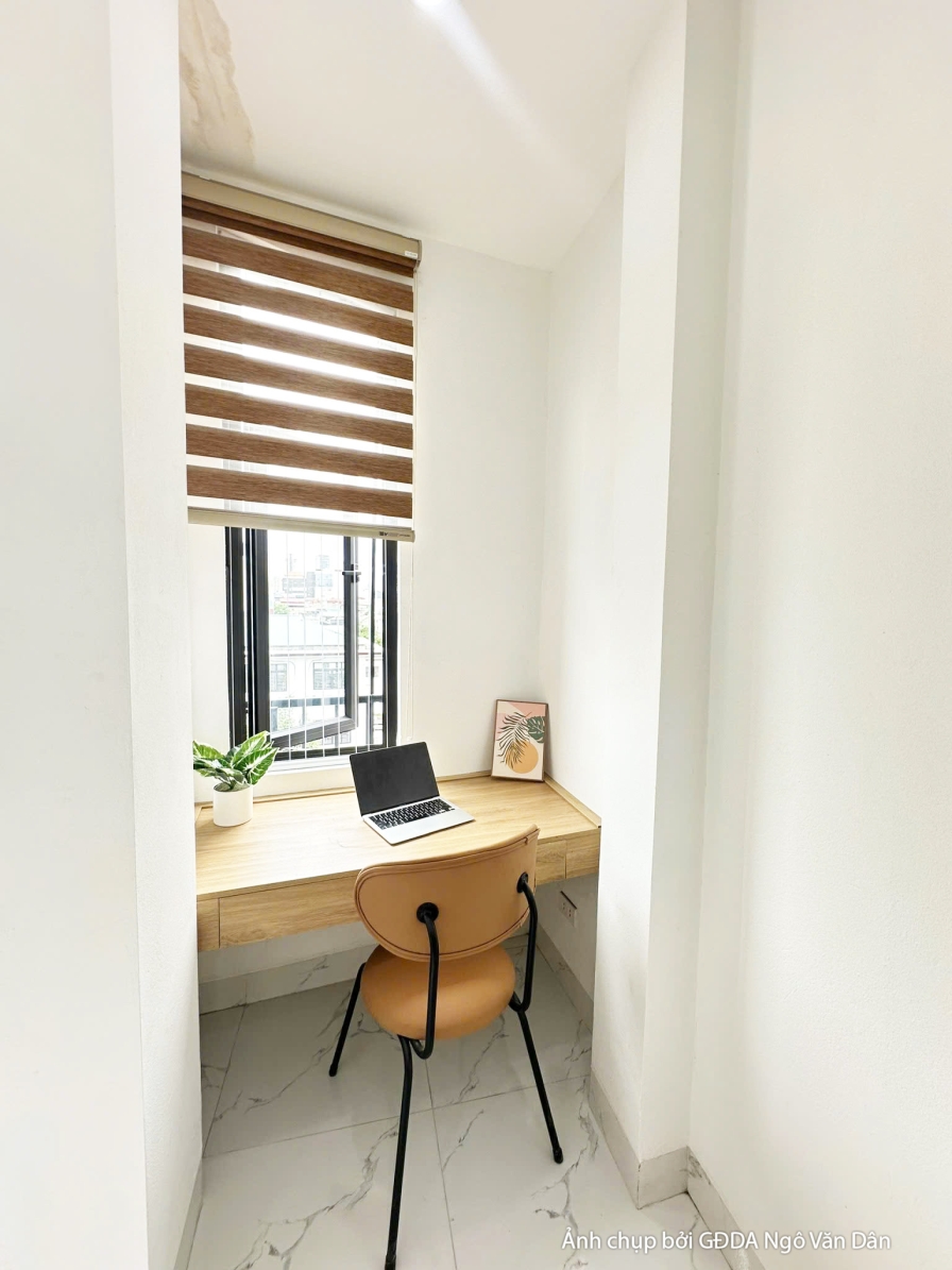 Góc làm việc nhỏ gọn trong phòng ngủ với bàn gỗ đơn giản, ghế và kệ trang trí. Cửa sổ lớn với rèm sáo gỗ cho phép ánh sáng…