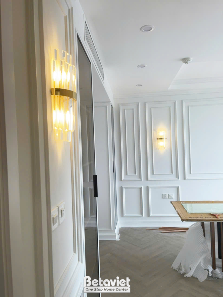 Chi tiết tường ốp phào chỉ trắng với đèn tường hiện đại, ánh sáng vàng dịu nhẹ, làm nổi bật các đường nét trang trí tinh x…