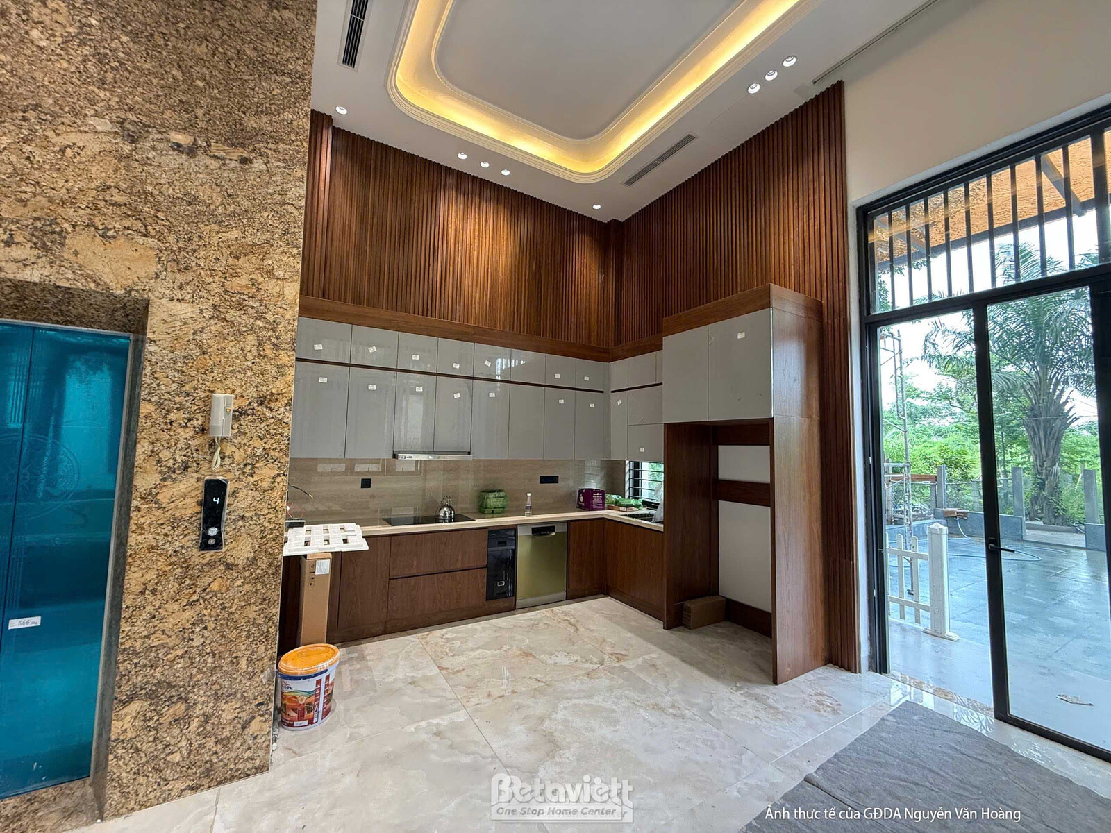 Không gian bếp đang trong giai đoạn lắp đặt hoàn thiện. Hệ tủ bếp chữ L màu nâu gỗ kết hợp cánh tủ sáng màu, mặt bếp ốp đá…