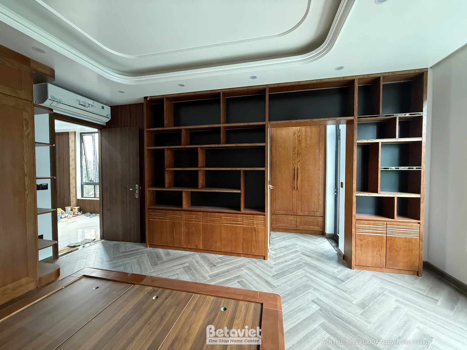 Phòng ngủ hoặc phòng làm việc đang lắp đặt nội thất gỗ. Hệ tủ sách lớn âm tường, giường gỗ và sàn xương cá. Trần thạch cao…