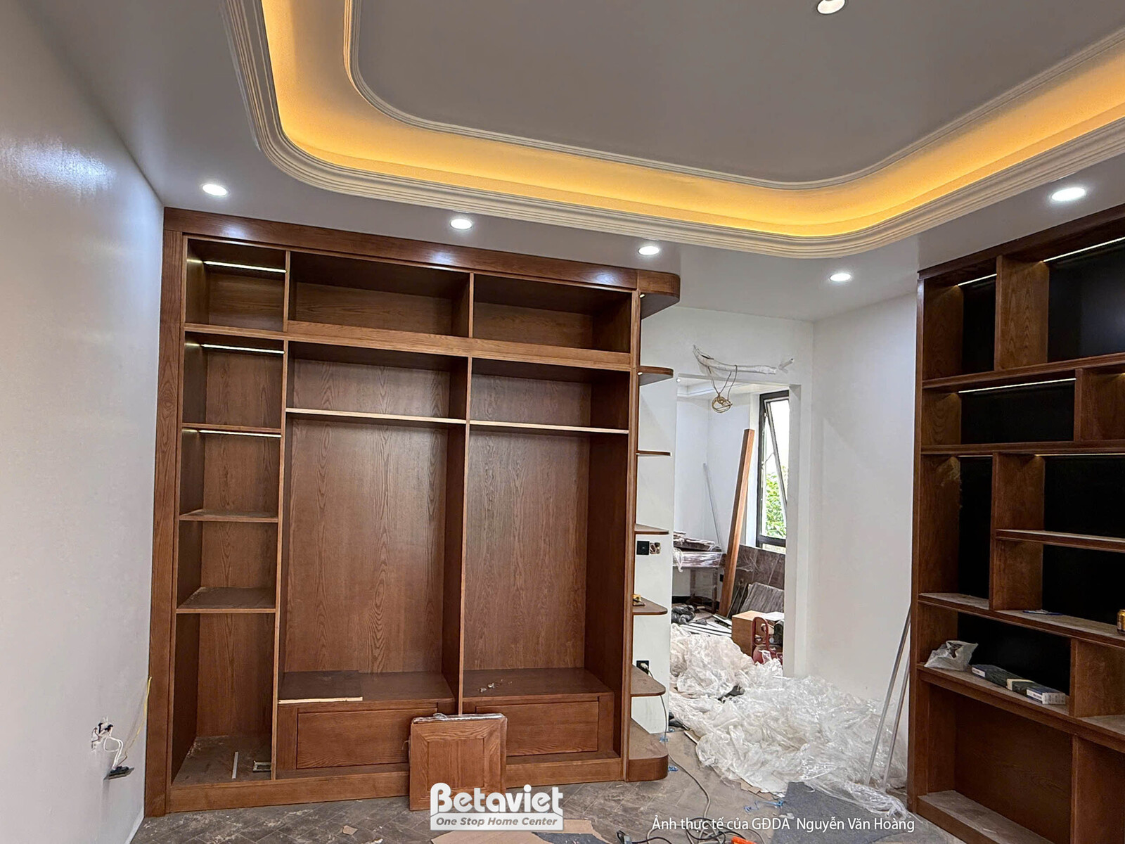Không gian phòng chức năng đang lắp đặt hệ tủ gỗ lớn âm tường. Tủ gỗ màu nâu trầm với nhiều ngăn kệ, trần thạch cao giật c…