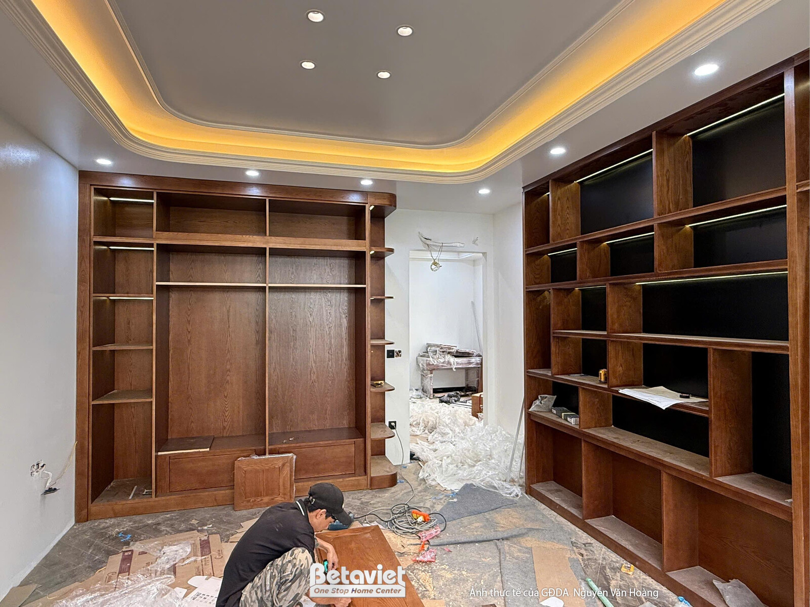 Một công nhân đang thi công lắp đặt hệ tủ gỗ âm tường trong phòng chức năng. Tủ gỗ màu nâu trầm, trần thạch cao giật cấp…
