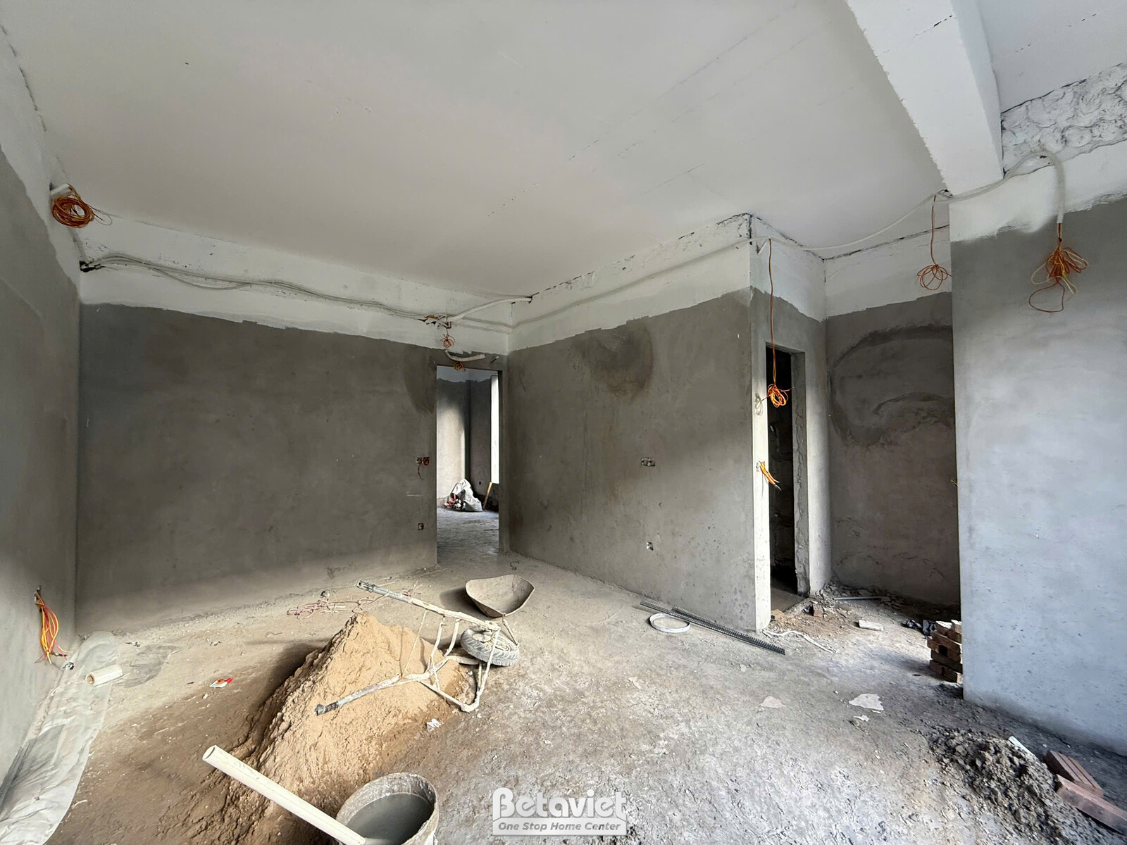 Một phòng chức năng đang trong giai đoạn phần thô, nhìn từ cửa vào. Sàn đất, tường bê tông chưa trát và các đường ống điện…