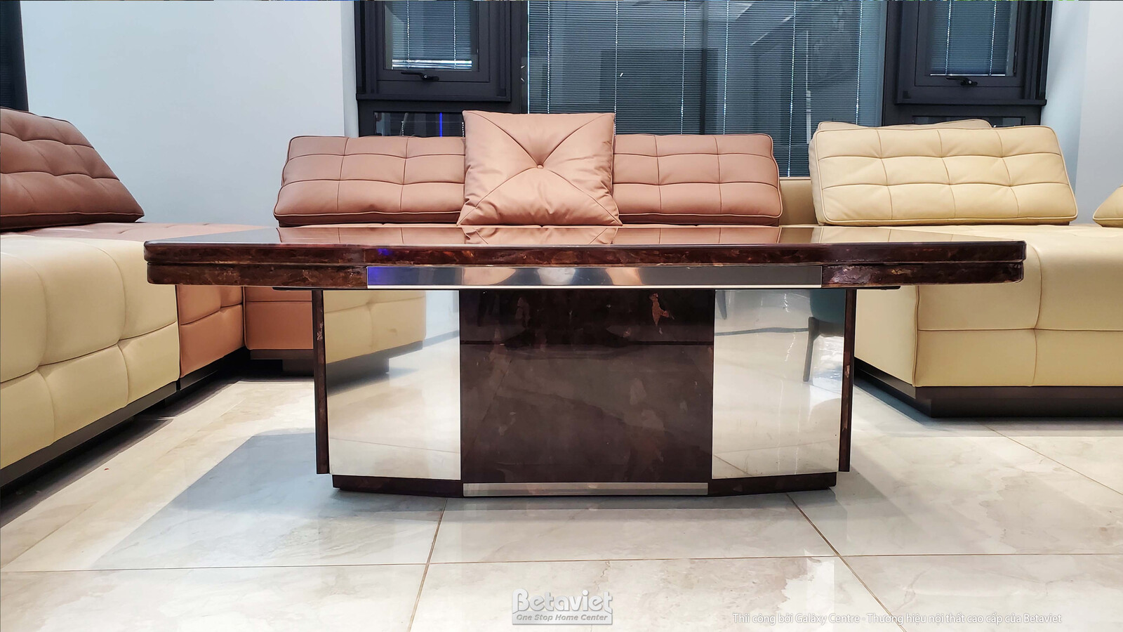 Cận cảnh bàn trà mặt đá vân nâu bóng với chân đế lớn, đặt giữa bộ sofa màu be và nâu nhạt. Thiết kế hiện đại, vật liệu cao…
