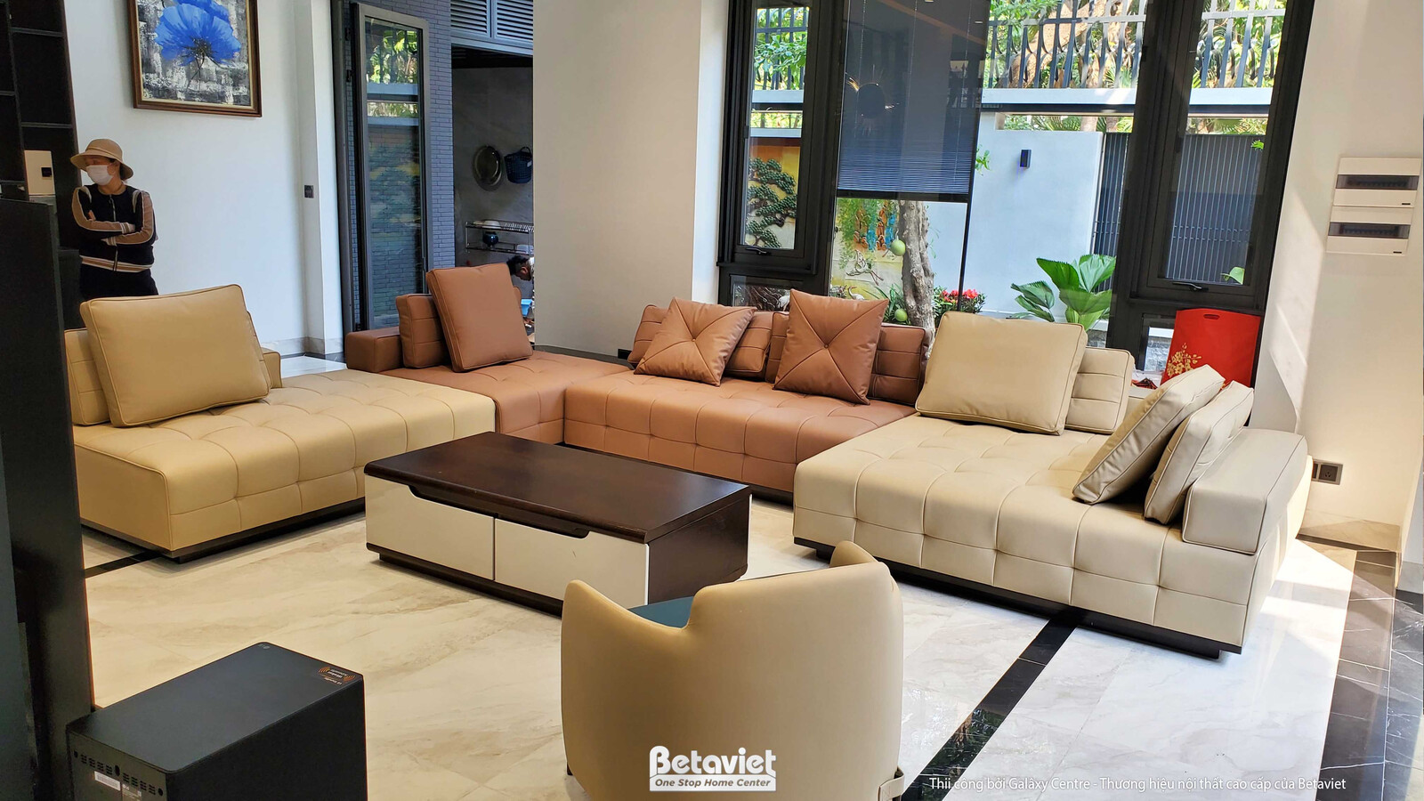 Góc phòng khách với bộ sofa chữ L màu be và nâu nhạt, bàn trà gỗ tối màu. Sàn lát đá vân sáng, không gian mở, tràn ngập án…